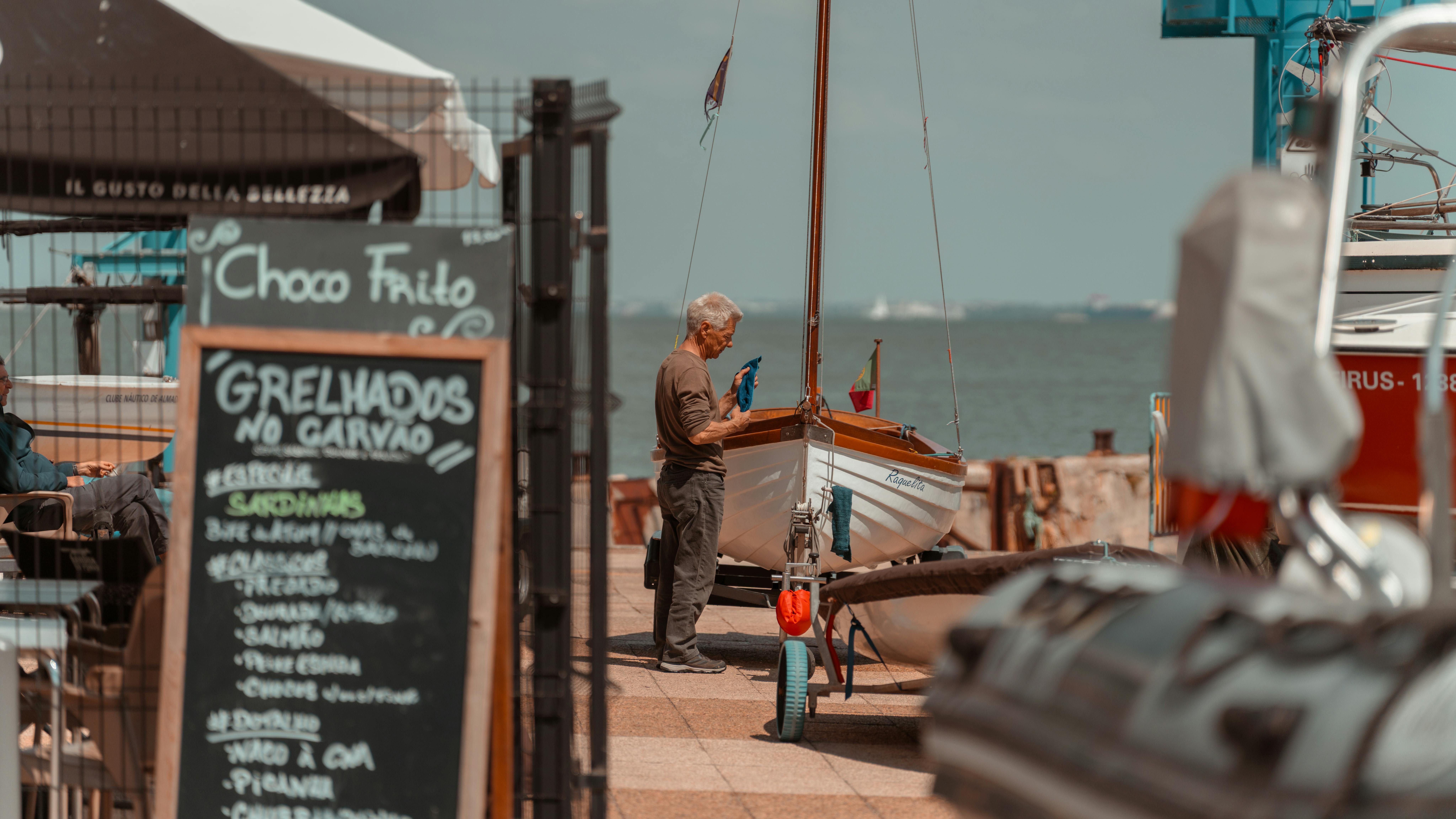 An older man works on a small wooden sailboat at a sunny Portuguese waterfront, with a seafood restaurant chalkboard menu in the foreground